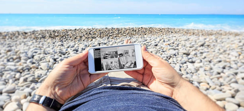 Egy ember a tengerparton a mobiljáról távoli biztonsági kamerákat néz